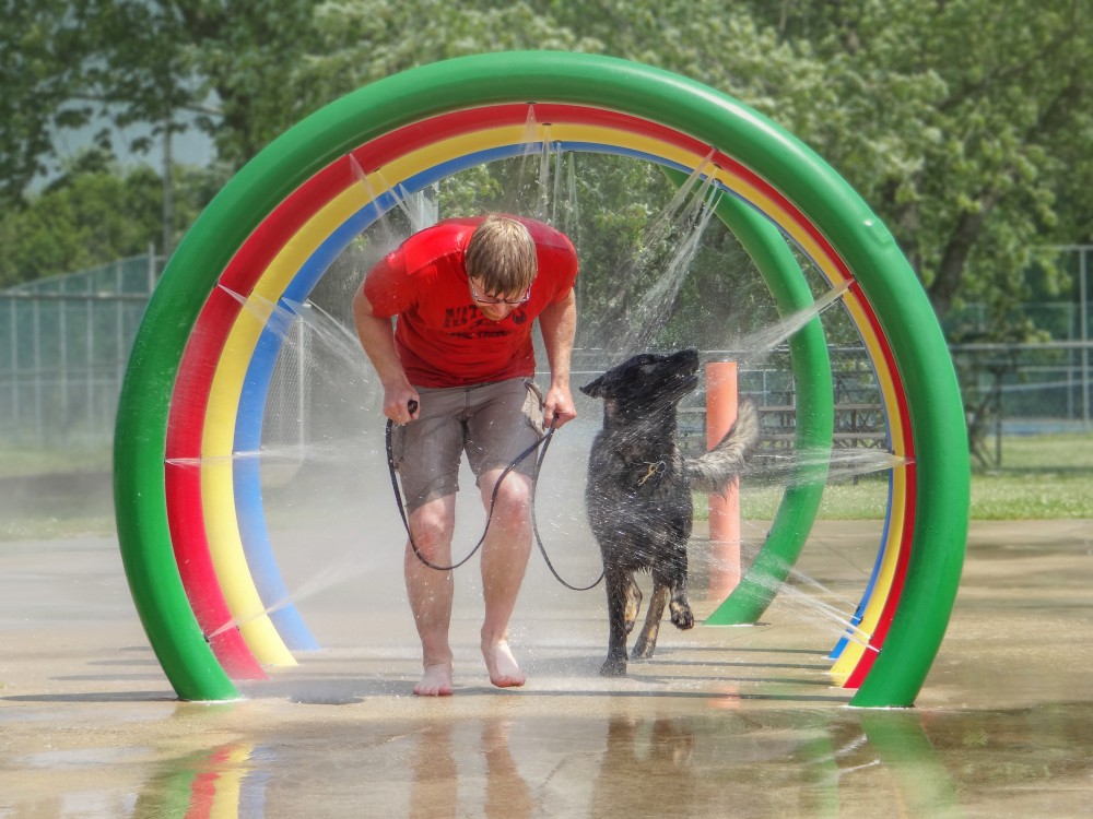 The photo shows a man and his dark sable German Shepherd Dog moving through a water spray feature at a park. Both are soaked. Martin, wearing an orange T-shirt and dark beige shorts, is bent forward as he moves through the spray. Bosco has his right paw raised horizontally and his muzzle turned directly into the water, clearly enjoying the blast. A colourful water feature arches over them, spraying water in multiple directions. Green trees and a park area are visible in the background.